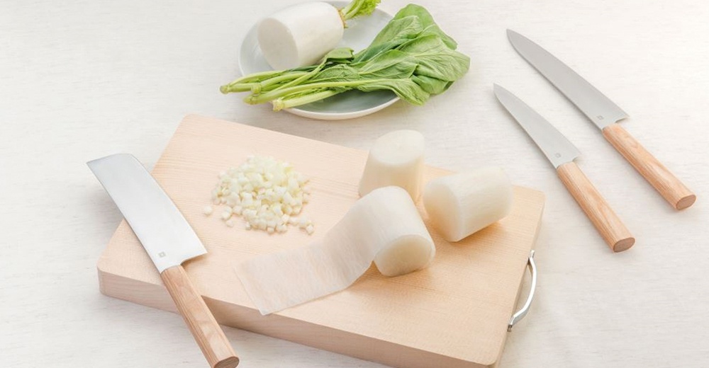 Yuri 萬用刀，配有木質手柄，刀刃為銀色不鏽鋼。圖片展示了一把萬用刀、兩把日式菜刀，以及切好的白蘿蔔塊、切碎的洋蔥和一株青菜，所有物品都放在木製砧板上。
