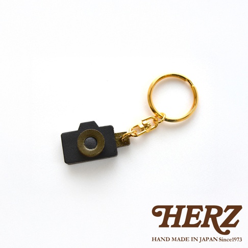 職人手作皮革相機造型鑰匙圈，黑色皮革製成，搭配復古金色鏡頭裝飾，連接金色鑰匙圈。