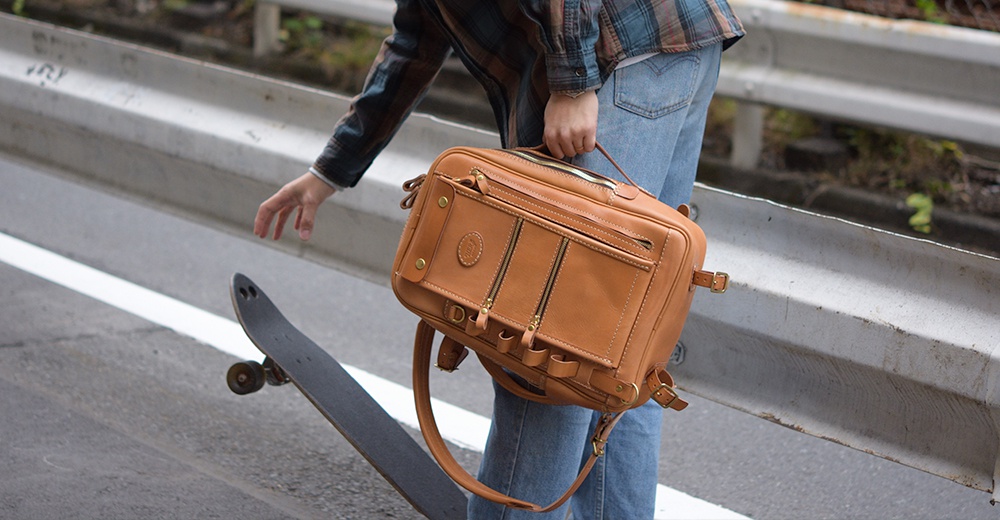 職人手作皮革兩用多功能後背包 R-66，採用棕褐色皮革製成，設有頂部提把、可拆卸肩帶、多個拉鍊隔層，以及金屬扣環和鉚釘裝飾。