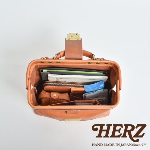 這是一款由日本HERZ品牌職人手作的皮革口金包，採用俯視視角呈現其內部空間。駝色皮革質感細緻，包內整齊收納了筆記本、錢包與各式生活小物，展現出俐落且實用的收納機能，包身結構堅固並配有金屬扣環，底部印有HERZ品牌商標與創立年份。