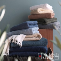 圖片中有一疊疊的毛巾，顏色從白色、米色、棕色到深藍色和綠色不等，毛巾堆疊在木架上，左側有一條米白色的毛巾，上面有流蘇，右下角有日本桃雪的標誌。