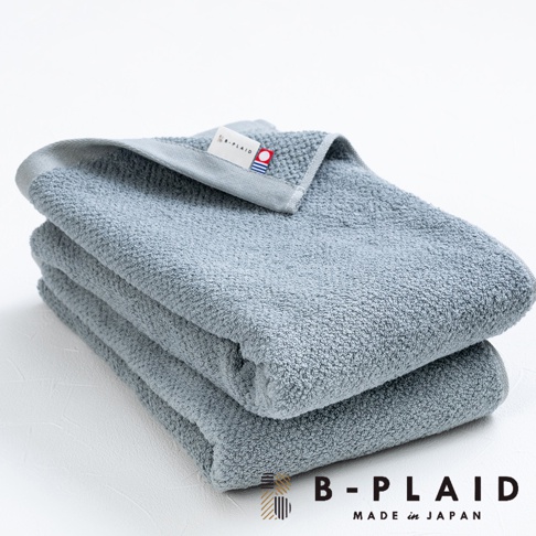堆疊的灰色薄手鱗紋浴巾，頂部一條帶有「B-PLAID」標誌的織標，材質柔軟，觸感細緻。