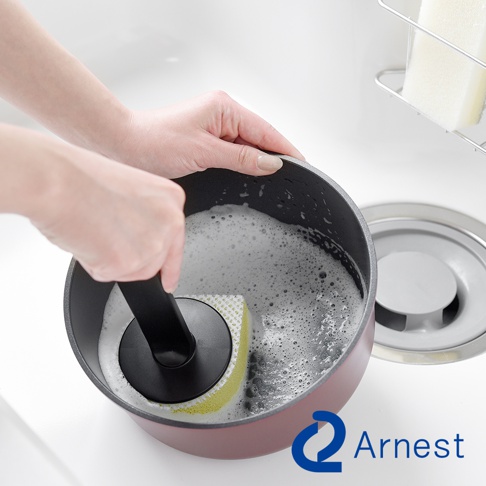 Arnest 品牌標誌下方，有人正在用帶有黃色海綿的黑色刷子清洗帶有泡沫的鍋子，鍋子放在水槽邊。