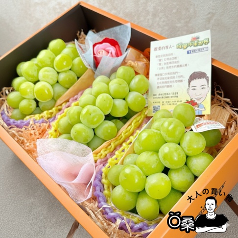 綠色日本麝香葡萄禮盒，內有三串飽滿的葡萄，搭配粉紅色和紅色花朵裝飾，以及印有卡通人物的卡片。