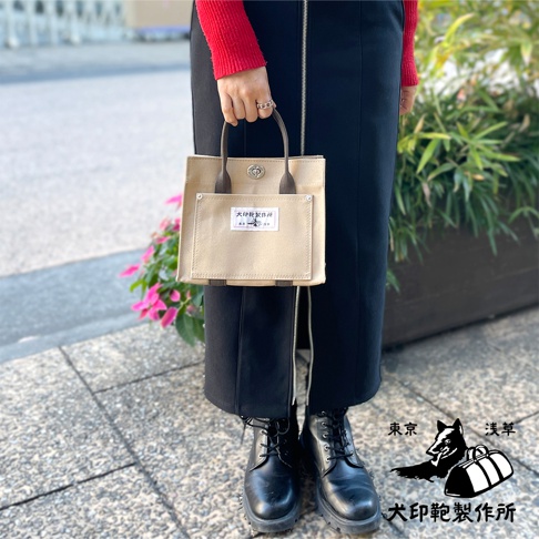 一手持淺米色純棉帆布迷你托特包，包身正面有一個同色系方形外袋，上方印有日文標籤。提把為深棕色。