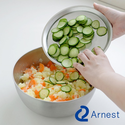 防刮不鏽鋼調理碗中裝有馬鈴薯和胡蘿蔔丁，一隻手正將切片的小黃瓜從不鏽鋼盤中倒入碗裡。