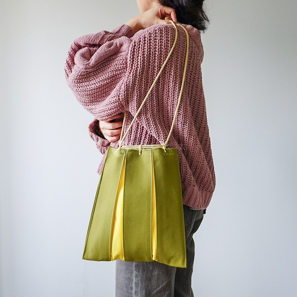 一位女性側身站立，背著一個京都KOSHO品牌的ougi帆布信玄袋。該手袋外層為橄欖綠色帆布，拼接鮮黃色布料，呈現出摺扇般的立體皺褶設計。袋口配有淺金色編織提繩，整體風格簡約且具有日式雅緻感，搭配粉紫色粗針織毛衣與灰色牛仔褲。