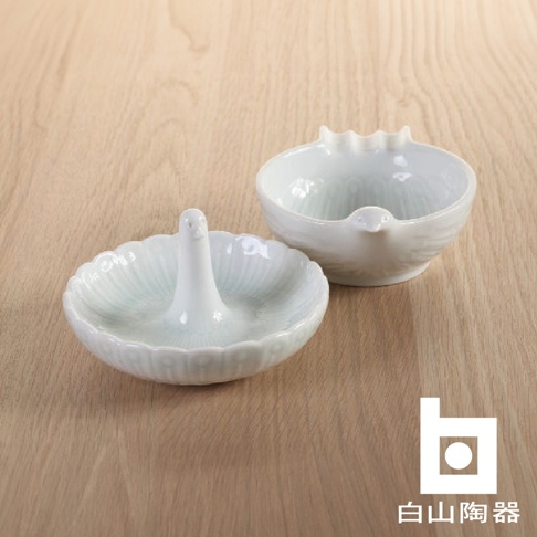 兩個白色陶瓷小碟，其中一個碟中央有直立的鳥形支架，另一個碟則有鳥嘴造型，兩者均有精緻的紋理裝飾，置於木質背景上。