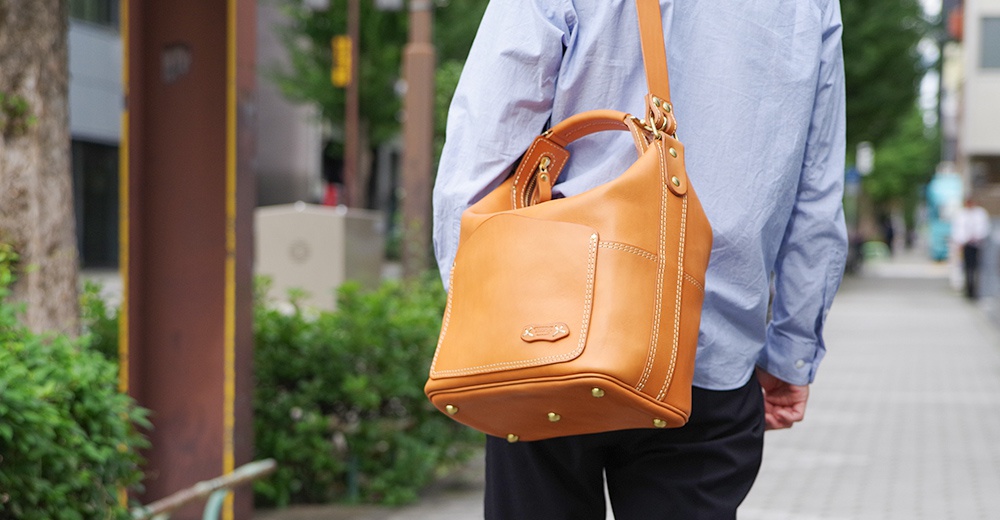 一個棕褐色皮革製成的厚實單肩波士頓包，包身有明顯的縫線裝飾，正面有一個翻蓋口袋，底部有金屬腳釘。