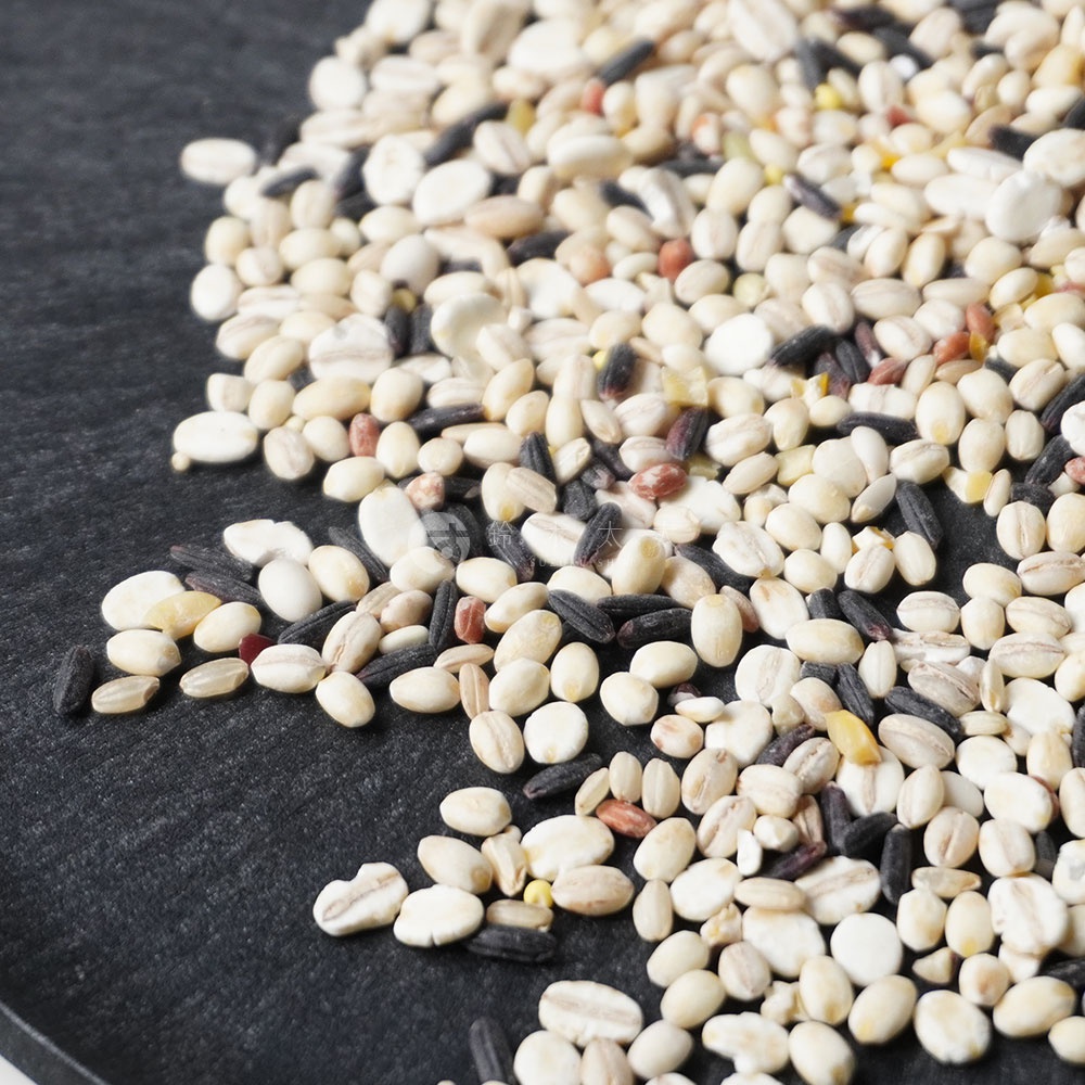 一堆由多種穀物組成的混合米，包含白色、黑色、淺棕色和紅色的穀粒，以特寫方式散落在深色背景上。