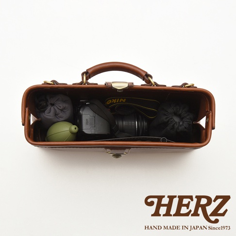 HERZ 職人手作皮革橫長形杜勒斯單肩包，內部裝有相機、鏡頭及其他配件，上方為提把，品牌標誌於下方。