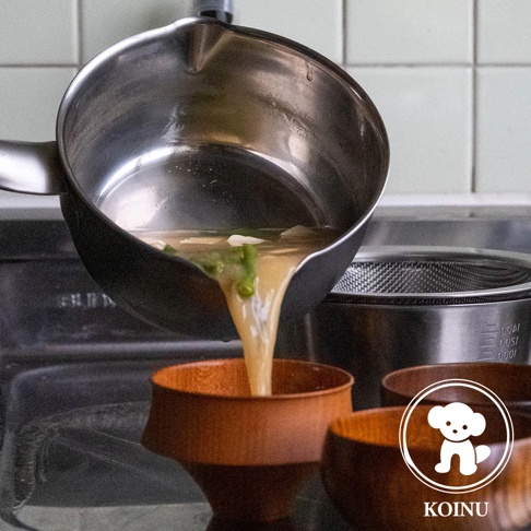 三層不鏽鋼雪平鍋傾斜，鍋內湯汁正倒入木碗中。鍋身為銀色不鏽鋼材質，外側為黑色塗層，鍋緣有導流設計。背景為廚房環境，可見濾網和瓷磚牆面。