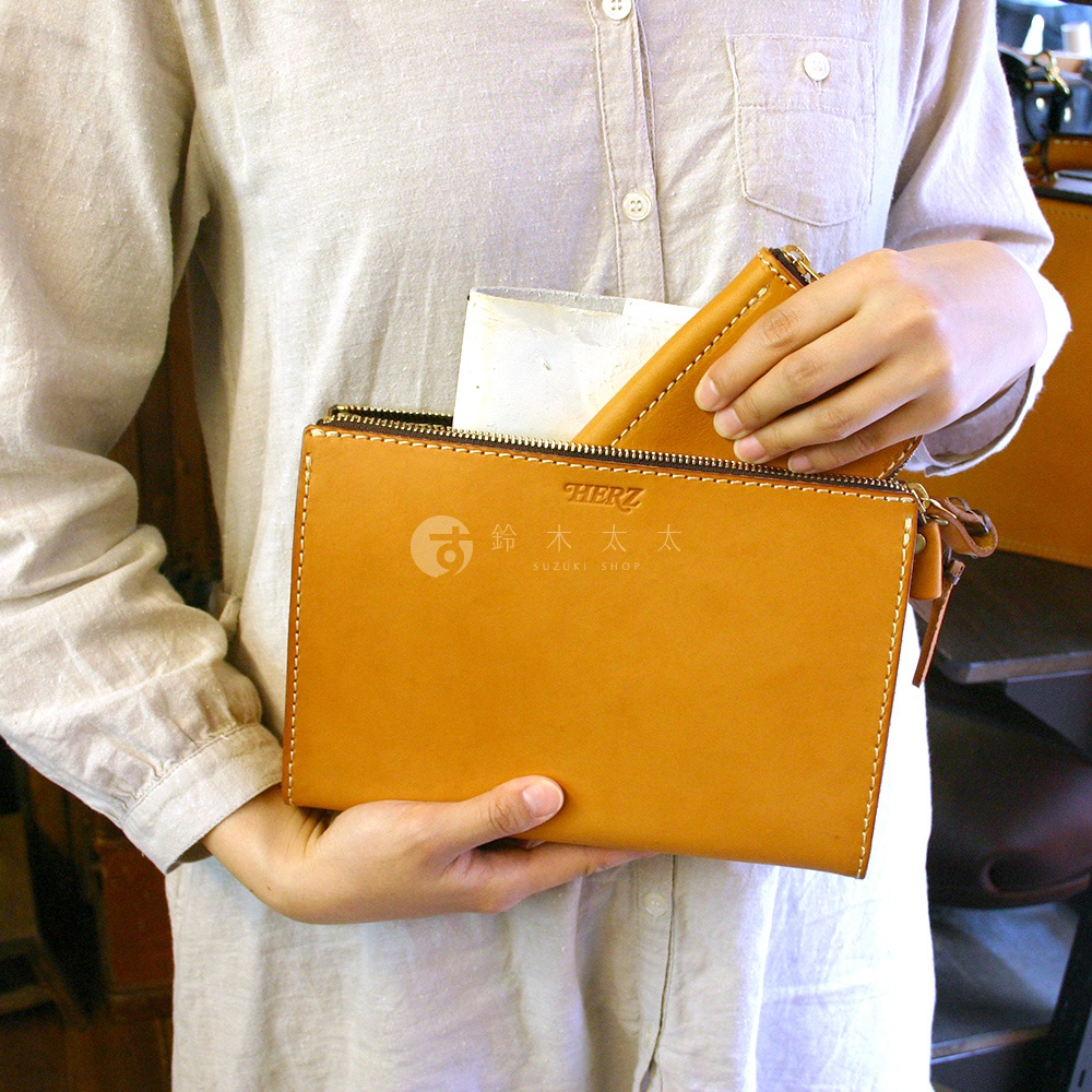 焦糖色皮革手拿包，配有拉鍊開口和品牌標誌，由雙手持握，展現簡潔的設計。