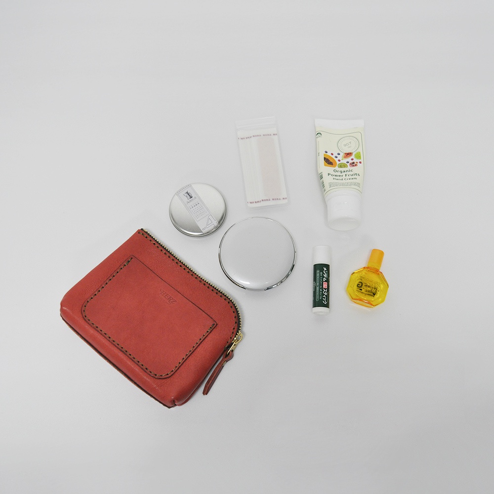 職人手作皮革小物收納包，橘紅色，展示於白色背景上，旁邊擺放著手部乳液、護唇膏、眼藥水、小圓罐及透明包裝的OK繃。