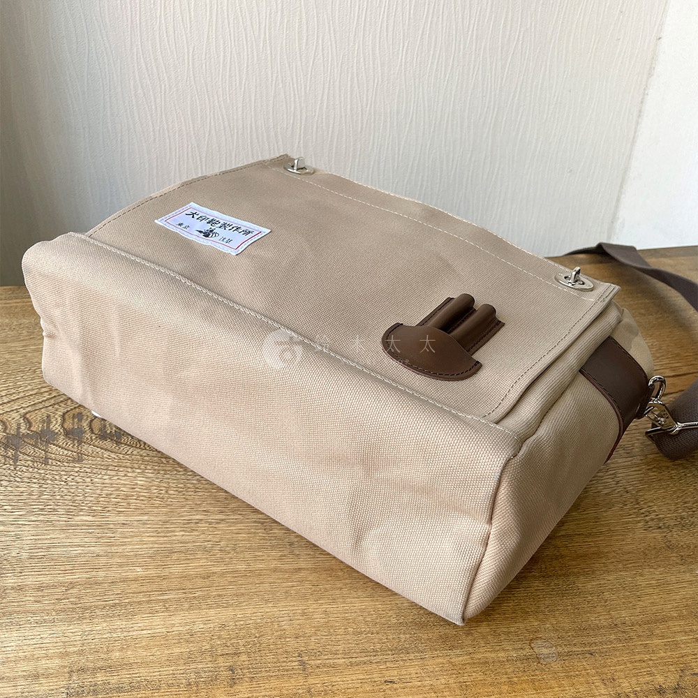 卡其色純綿帆布製成的橫式側背包，帶有深棕色皮革裝飾，包蓋上有品牌標籤及金屬扣環。