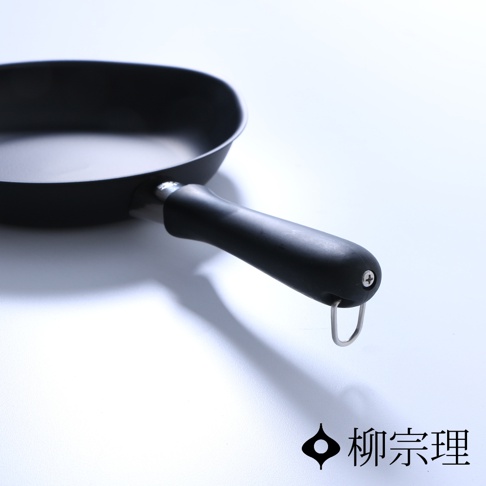 一個深灰色岩紋質感的柳宗理單手鐵鍋，採用霧面黑色塗層，手柄末端設有金屬懸掛扣環與固定螺絲，整體設計簡約現代，右下角標示有柳宗理品牌名稱。
