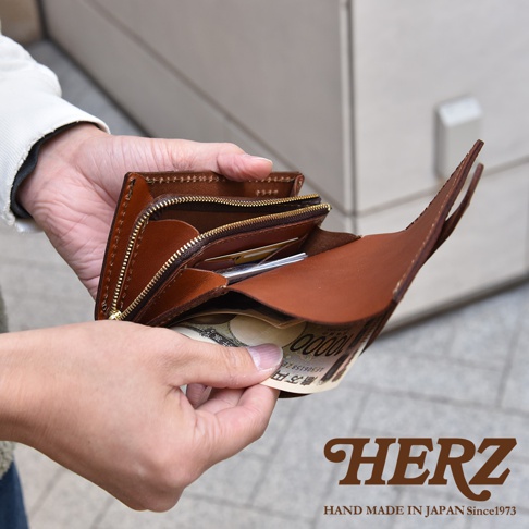 這張特寫照片展示了一位消費者正手持一個日本HERZ職人手作的棕色皮革中型皮夾，皮夾內夾著一張萬元面額的日圓紙鈔，皮革質感細膩且縫線精緻，皮夾設計包含拉鍊零錢層與多個收納空間，呈現出經典耐用的手工藝風格，圖片右下方印有品牌標誌與手工製造年份。