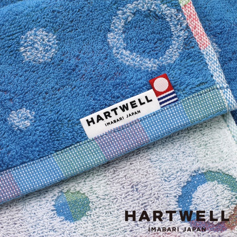 這是一張近距離拍攝的 Hartwell 今治毛巾局部特寫，畫面呈現深藍色與淺藍色兩條毛巾重疊的樣貌，毛巾表面飾有圓點花紋，邊緣處帶有細緻的織紋格狀裝飾。上方深藍色毛巾縫製著白色長方形標籤，上面印有黑色 Hartwell Imabari Japan 字樣，標籤一角裝飾著紅、藍相間的圓形圖標，展現細緻的毛巾織造工藝與品牌質感。