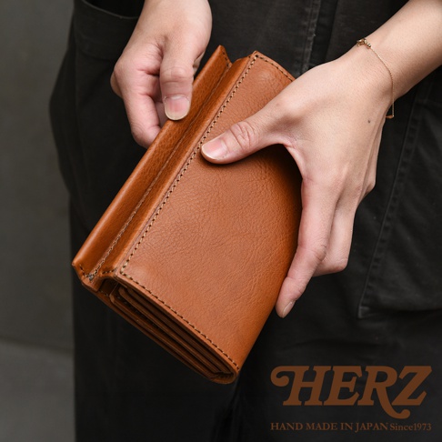 一雙手正在持拿一個棕色皮革長夾，該長夾呈現出厚實溫潤的質感，皮革邊緣有細緻的手縫線跡，整體為Organ系列的職人手作設計。畫面右下角有HERZ品牌標誌與手工製造年份說明。