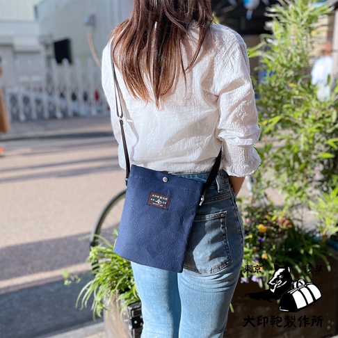 深藍色燈芯絨材質的平板收納側背包，配有黑色細肩帶，包身有品牌標籤，由一位身穿白色刺繡襯衫和牛仔褲的女性背在身上，背景為戶外街道景觀。