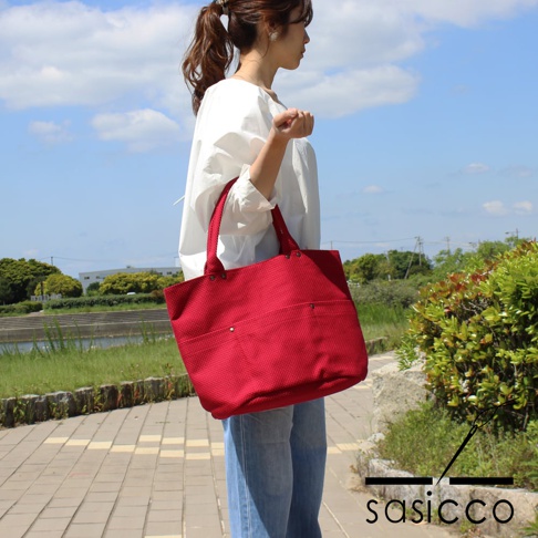 一位女性身穿白色上衣和牛仔褲，肩背一個紅色三河木綿刺子織大開口托特包，包包有兩個外部口袋，材質表面有細小的格紋織紋。