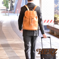 一位男士身穿西裝，背著一個棕褐色的皮革單扣束口後背包，背包有抽繩和金屬扣環。