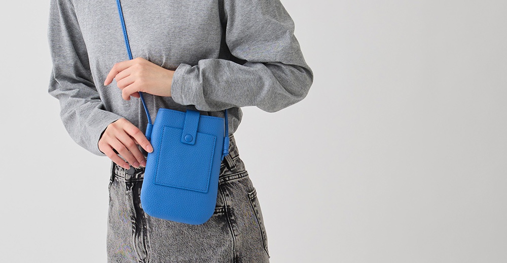一個穿著灰色長袖上衣和牛仔褲的人，右肩上斜背著一個藍色皮革手機包。