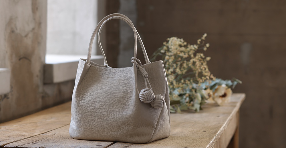 itten-itten Neru 義大利皮革點結吊飾中型托特包，採用細緻顆粒感的淺灰色義大利皮革製成，配有品牌標誌壓紋及可愛的點結皮革吊飾，風格簡約優雅。