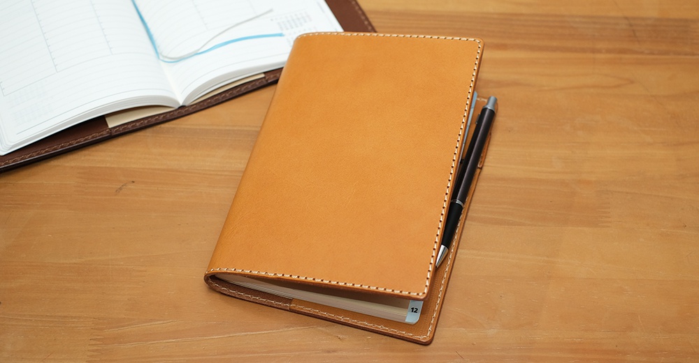 淺棕色皮革B6記事本書套，採用車縫線裝飾，搭配黑色鋼筆，放置於木質桌面。