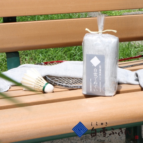 紀州備長炭紗布長巾，白色，材質為紗布，包裝內含一條長巾，旁邊擺放著羽毛球拍和羽毛球，皆放在木製長椅上。