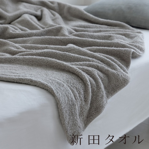 一張以近距離視角拍攝的灰色毛巾材質單人被，柔軟的織物自然垂落在白色的床鋪上，呈現出細膩的絨毛質感與皺褶紋理，右下角印有深褐色的新田タオル字樣。