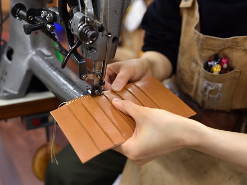 職人手作皮革多用途收納長夾 (KB-5)，展示製作過程中，雙手正在工業縫紉機上縫製淺棕色皮革。皮革呈現多層次摺疊設計，縫線清晰可見。