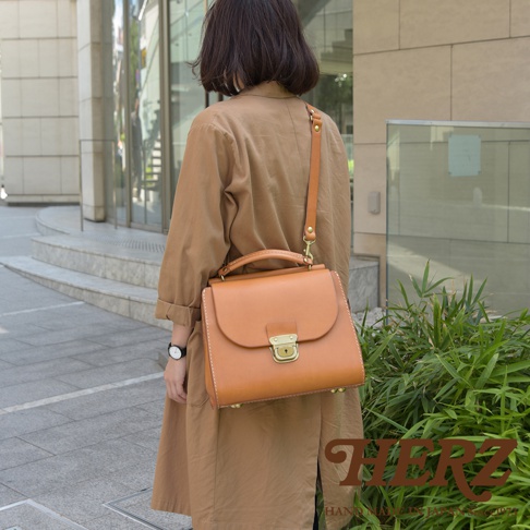 日本HERZ手工皮革屋瓦風兩用單肩包，由一位女士背著，包款為淺棕色硬挺皮革，搭配金色鎖扣，呈現簡約復古風格。