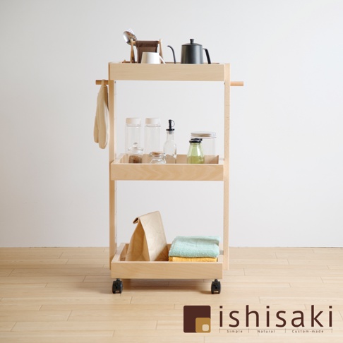 淺色木製三層收納推車，頂層有黑色水壺、白色杯子、餐具組和收納架；中層擺放數個透明儲物罐，其中包含調味料和綠色液體；底層有紙袋及疊放的藍綠色與黃色毛巾，推車下方裝有四個黑色輪子。