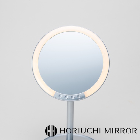 銀色圓形LED化妝鏡，鏡框周圍有暖白色LED燈光，鏡面下方有三個控制按鈕，產品立於銀色支架上，背景為淺灰色。