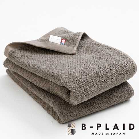 疊放的 EVE 今治製大浴巾，呈現深棕色，表面有細緻的鱗紋織法，質料厚實，一角的織標標示著品牌名稱「B-PLAID」及「MADE in JAPAN」。