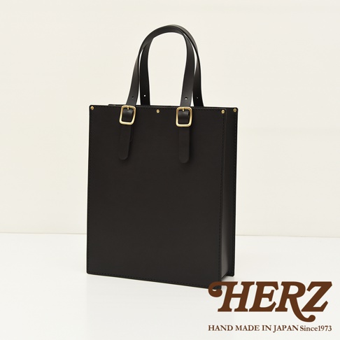 黑色皮革商務用紙袋托特包，配有兩條皮革提把，提把下方有金色搭扣裝飾。包款整體設計簡潔，線條分明，呈現出經典的風格。