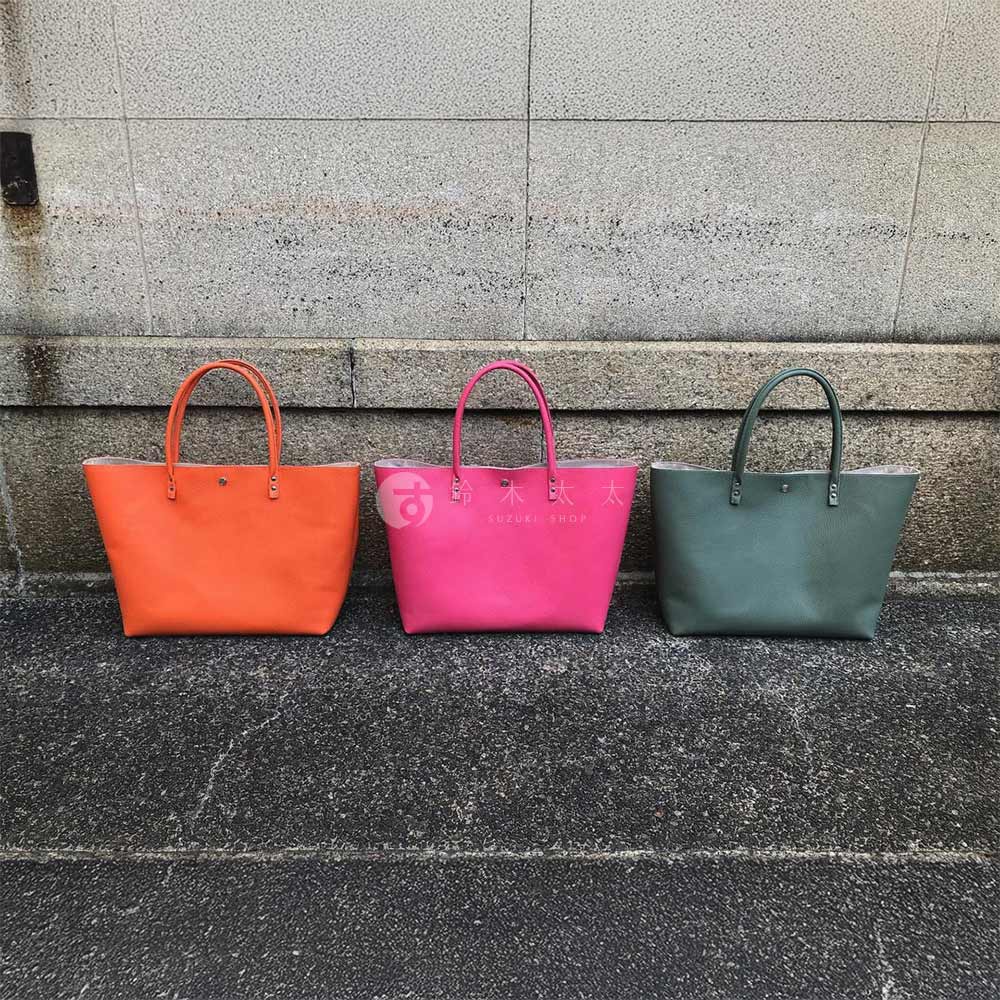 三款京都職人手作托特包，分別為亮橘色、桃粉色及墨綠色，皆為光滑皮革材質，簡約設計，配有提把。
