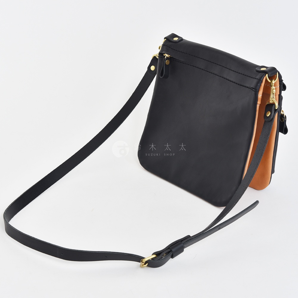 職人手作皮革翻轉雙色直式斜背包，主體為黑色皮革，側邊露出棕色皮革內襯，搭配金色五金配件與黑色皮質斜背帶，整體風格簡約。