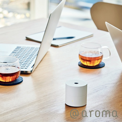 白色圓柱形隨身精油香氛機，頂部有圓孔，側面有黃色指示燈，放置在木質桌面上，旁邊有筆記本、筆、筆記型電腦和兩杯茶。