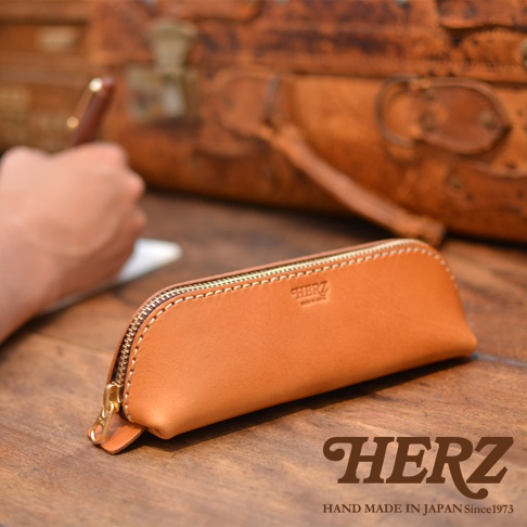 職人手作皮革三角收納筆袋，顏色為自然的棕褐色，材質為皮革，配有金色拉鍊和同色系縫線，袋身印有「HERZ」品牌標誌。