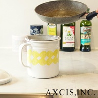 白色琺瑯濾油壺，壺身帶有黃色花紋，頂部有銀色濾網。畫面背景為廚房檯面，有油瓶和鍋子。