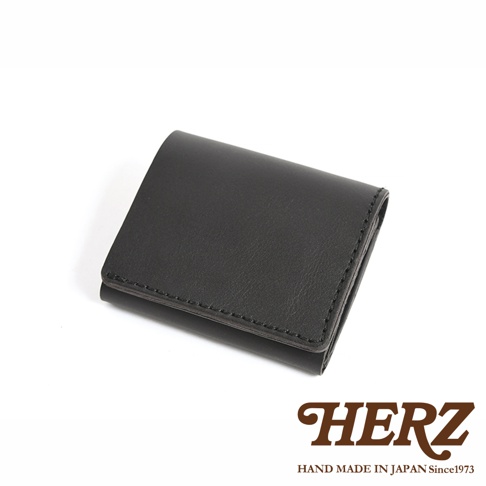 日本HERZ職人手作皮革雙折迷你短夾，呈現黑色霧面質感，皮件邊緣有細緻的手工縫線，整體外觀簡約素雅，右下角印有棕色的HERZ品牌商標與日本手工製造年份資訊。