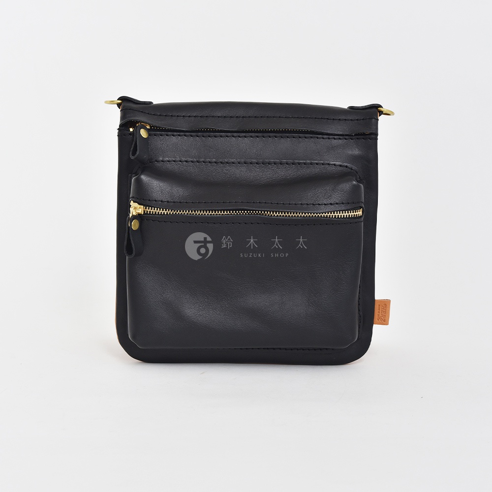黑色皮革翻轉直式斜背包，設有兩個拉鍊前袋，搭配金色拉鍊和金屬扣環。包包側面有品牌標籤。