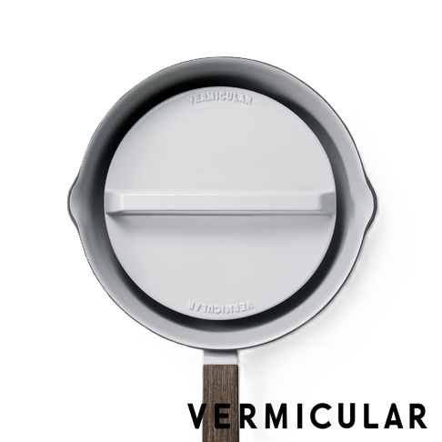 俯視視角展示一個 Vermicular 品牌的灰色琺瑯鑄鐵雪平鍋專用落蓋，表面帶有品牌刻字與一條橫向提把，搭配質感木紋手把的鍋身邊緣，整體設計簡約現代。