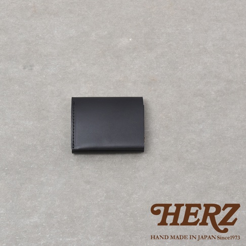 日本職人品牌HERZ Organ系列黑色手工皮革雙折迷你短夾，以單片義大利Minerva Liscio皮革製成，表面呈現極簡俐落的霧面質感，右下角印有品牌標誌，於淺灰色背景下呈現出精緻的職人手作風格。