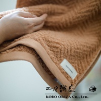 一隻手輕柔地觸碰著一件淺棕色、具有人字紋理的有機棉浴巾。浴巾邊緣有著米色的滾邊，上面縫有白色的小標籤。