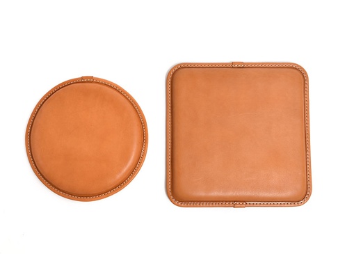 兩個職人手作皮革坐墊，一個為圓形，另一個為方形。兩者皆為淺棕色皮革材質，邊緣以米白色縫線包邊。