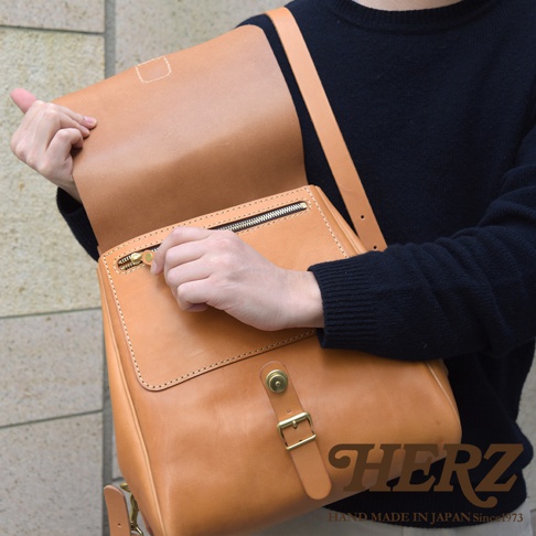 職人手作皮革屋瓦風後背包，採用天然皮革材質，呈現溫潤的淺棕色調。包款設有拉鍊主袋、前袋及金屬釦環，搭配寬版肩帶，設計簡潔實用。