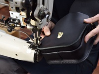 職人手作黑色皮革翻蓋迷你單肩包，包身上有金色旋鎖扣，正在由工業縫紉機縫製中。